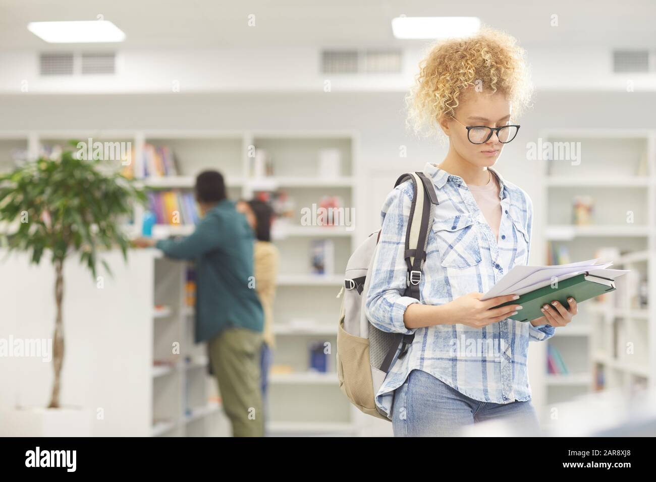 Girovita ritratto di donna bionda lettura libro mentre in piedi in biblioteca college, copia spazio Foto Stock