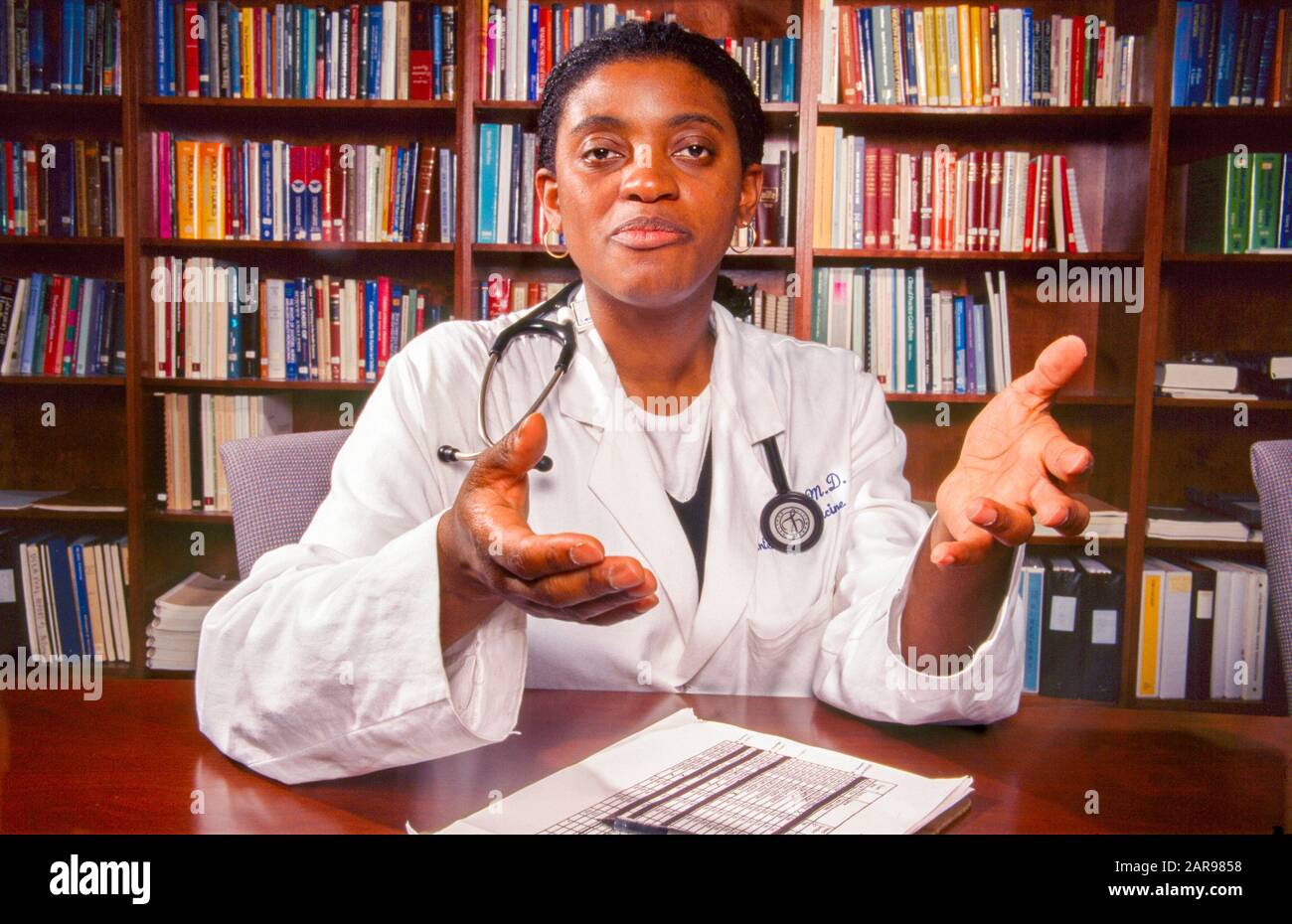 Indossando il suo cappotto bianco e con uno stetoscopio intorno alle sue spalle, un medico femminile afroamericano offre una spiegazione alla domanda di un paziente nel suo ufficio di Los Angeles, CA, rivestito di libri. VERSIONE DEL MODELLO Foto Stock