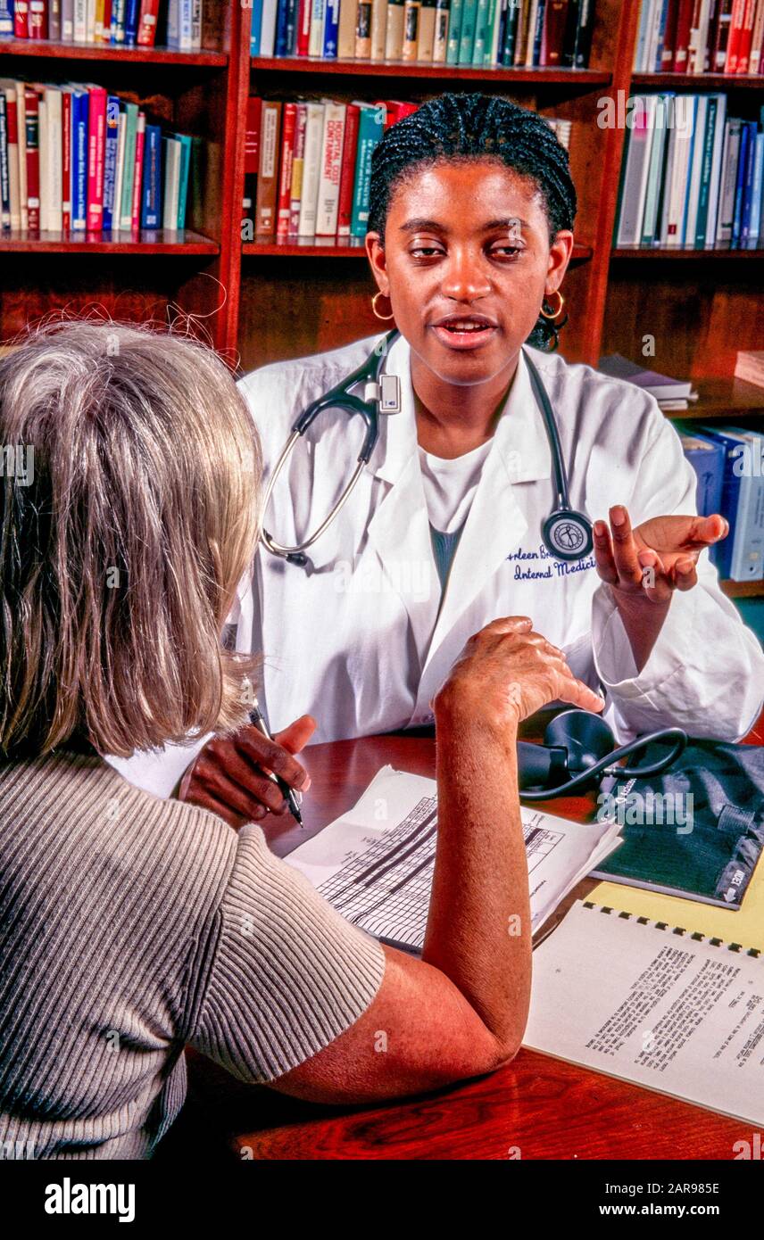 Indossando il suo cappotto bianco e con uno stetoscopio intorno alle sue spalle, un medico femminile afroamericano offre una spiegazione alla domanda di un paziente nel suo ufficio di Los Angeles, CA, rivestito di libri. Prendere nota del bracciale per la pressione sanguigna sulla scrivania. VERSIONE DEL MODELLO Foto Stock