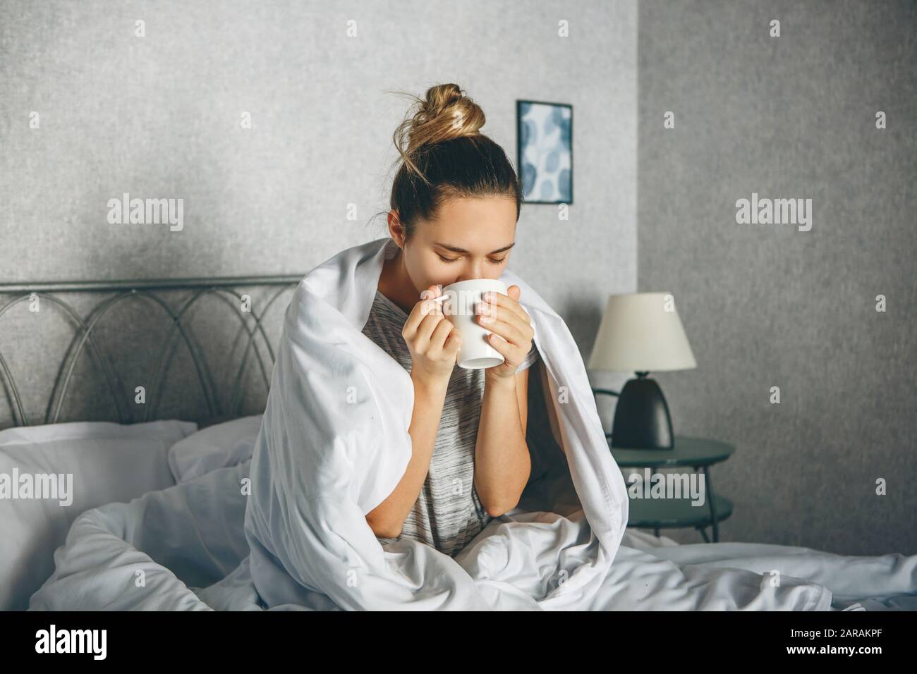 La ragazza beve un drink da una tazza nel letto avvolto in una coperta. Lei sta godendo o non è sveglio o malato. Foto Stock