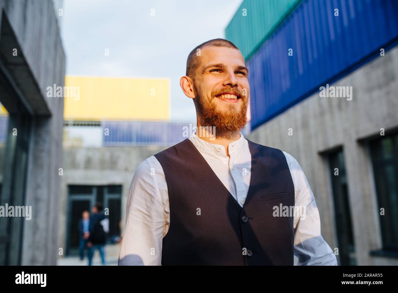 Felice uomo sorridente in una giacca senza maniche a piedi dalla strada stretta al tramonto Foto Stock