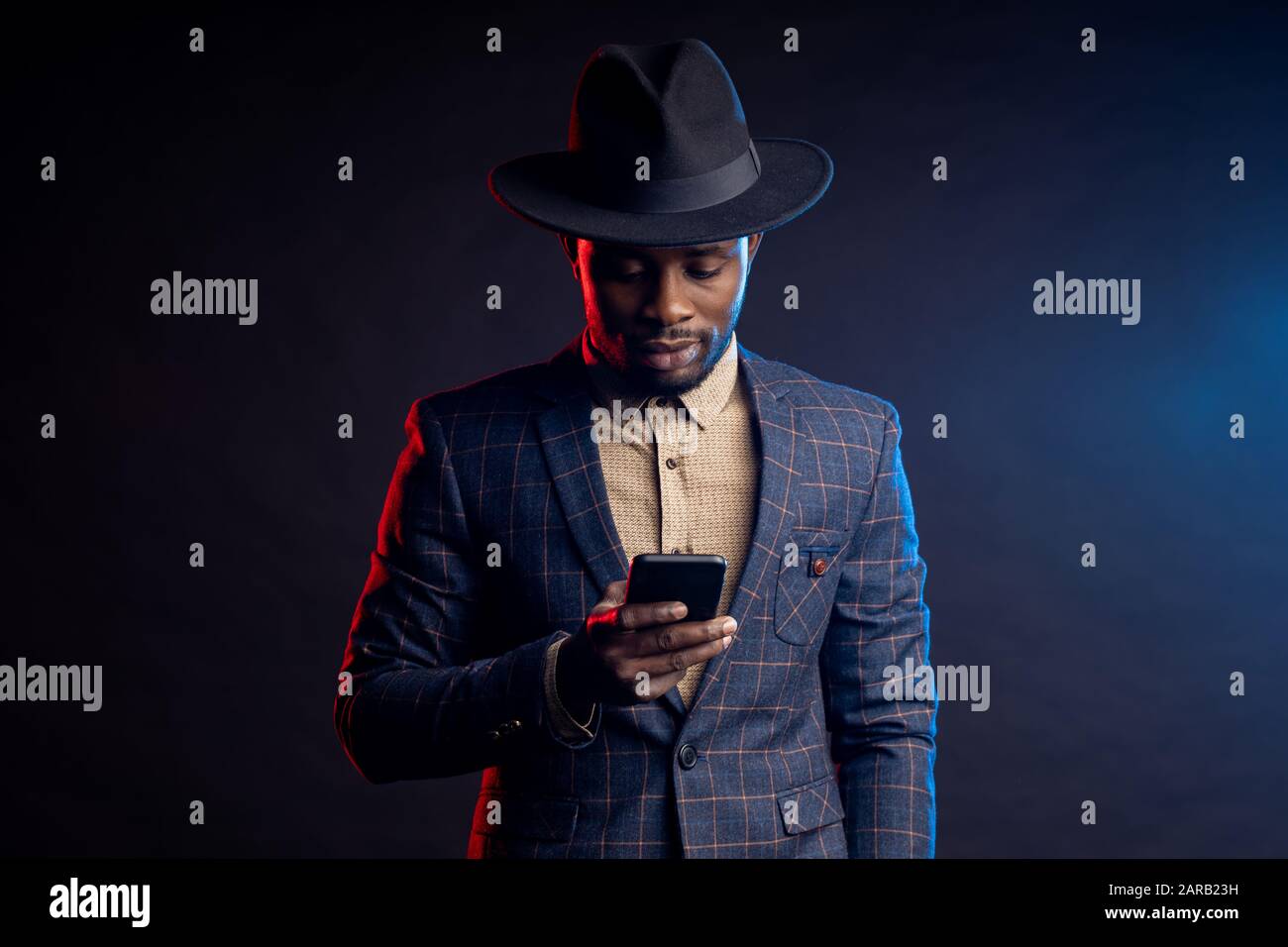 Girovita di sicuro elegante ragazzo africano, tenendo il telefono cellulare, guardando lo schermo, leggendo le notizie, chiacchierando con gli amici online, in piedi al buio Foto Stock