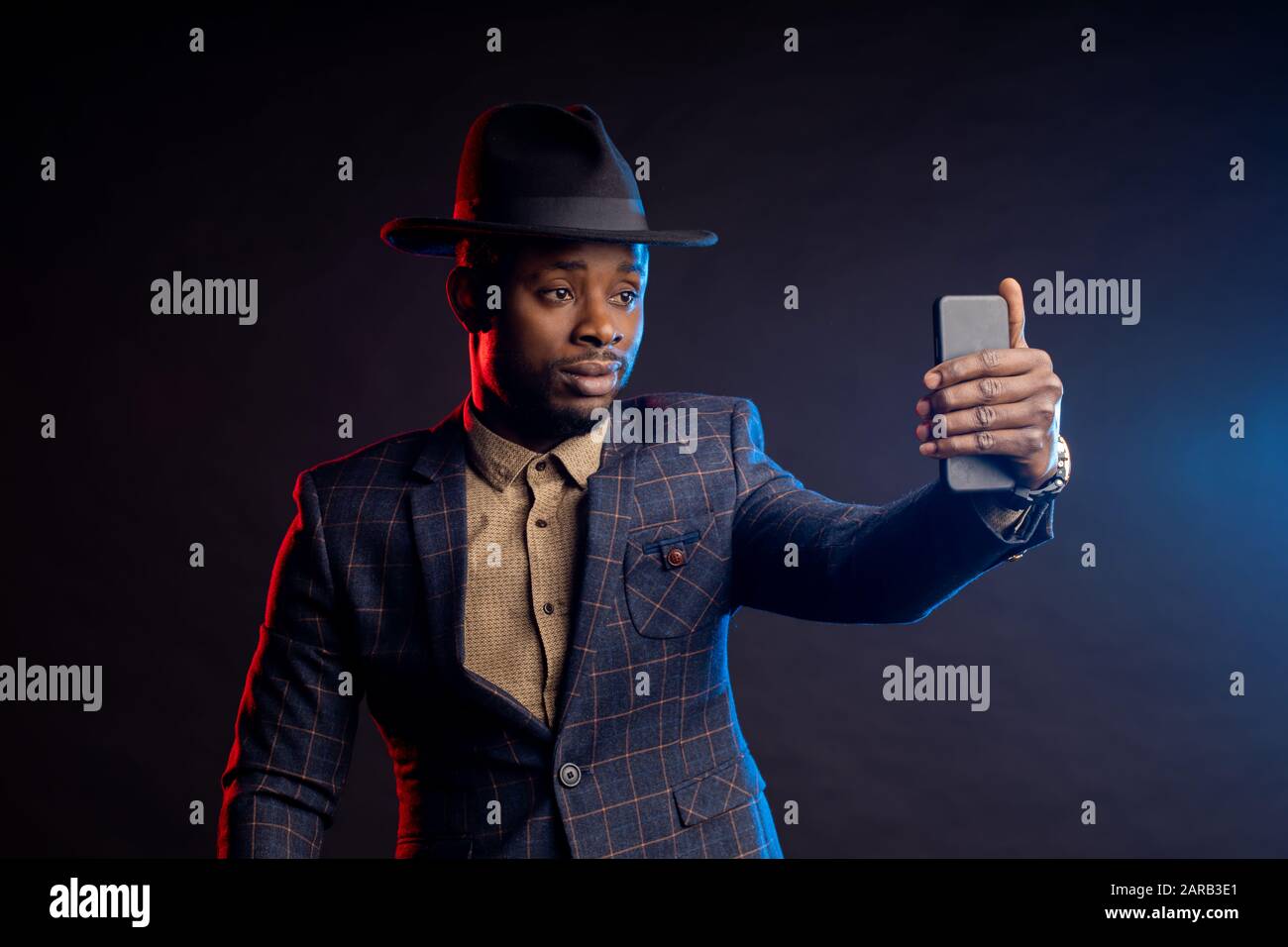 Un giovane afroamericano alla moda con bearded man che indossa un elegante abito a giacca, camicia, cappello, telefono cellulare, che porta selfie al buio, isolato in piedi Foto Stock