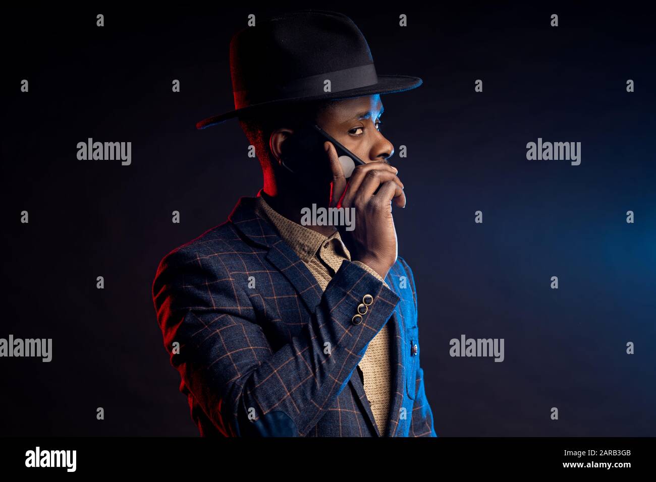 Scatto orizzontale di serio preoccupato giovane africano elegante vestito, cappello, parlando sul telefono cellulare, in piedi in buio su sfondo nero . Suc Foto Stock
