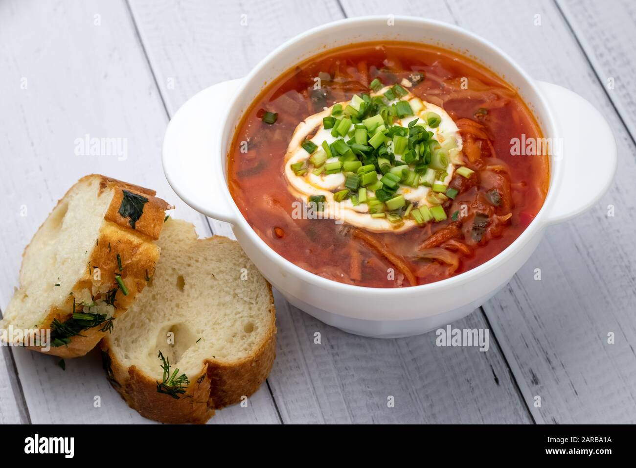 Borsct rosso ucraino con panna acida in ciotola bianca e pezzi di pane con aglio. Cucina russa concetto. Tavole di legno chiaro sfondo, tavolo Foto Stock