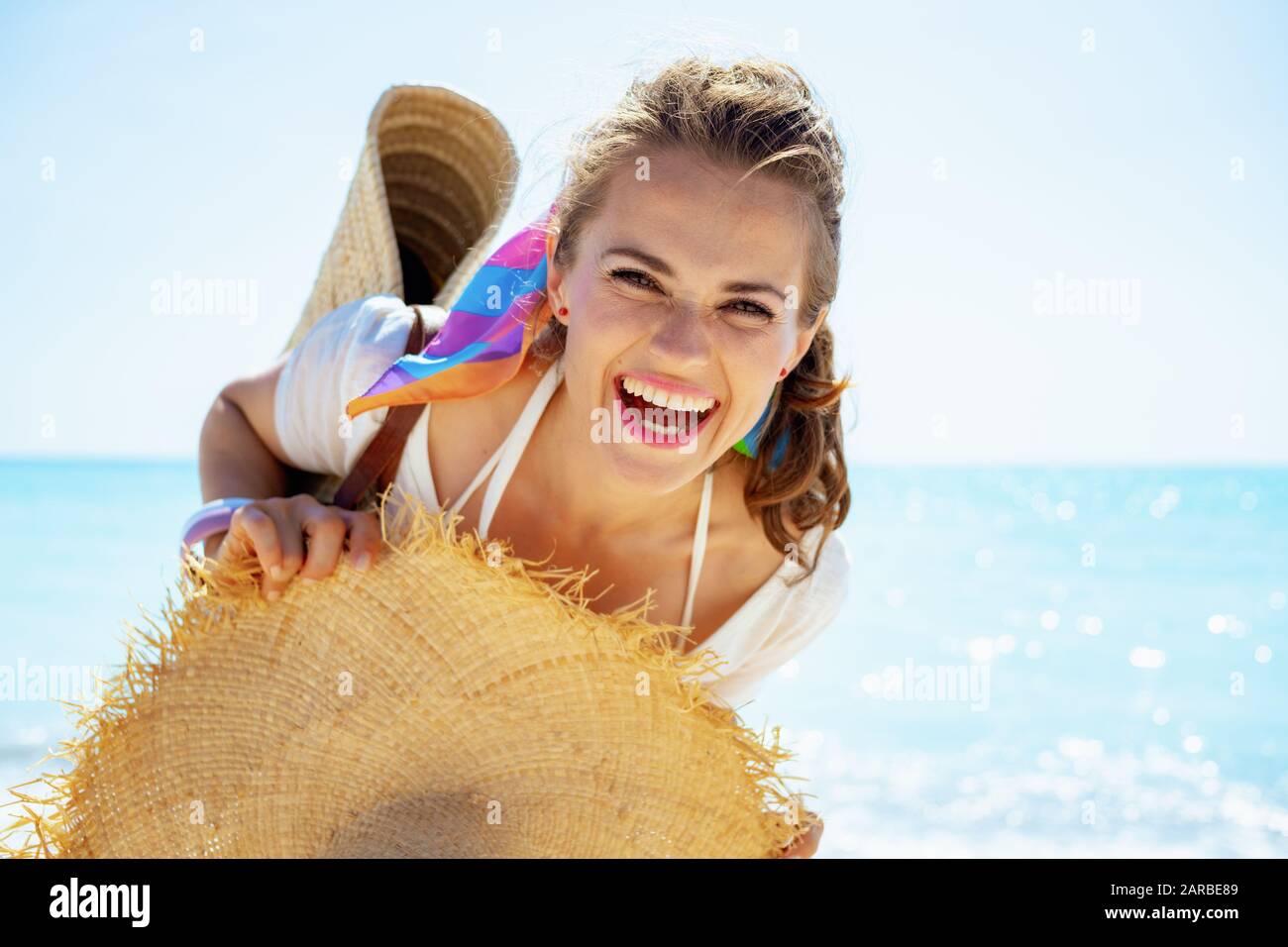 Ritratto di felice donna moderna di mezza età in maglietta bianca con borsa di paglia sulla spiaggia e grande cappello sulla spiaggia. Foto Stock