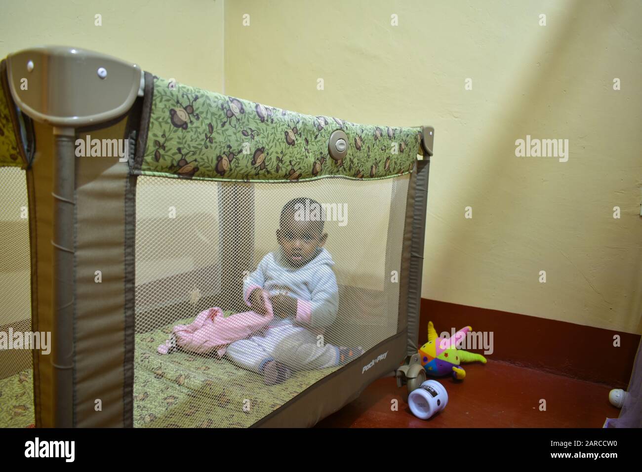 un bambino africano sta sbirciando attraverso la rete mentre gioca in un pack graco e gioca in un cortile. è adatto per viaggiare e può servire come letto Foto Stock