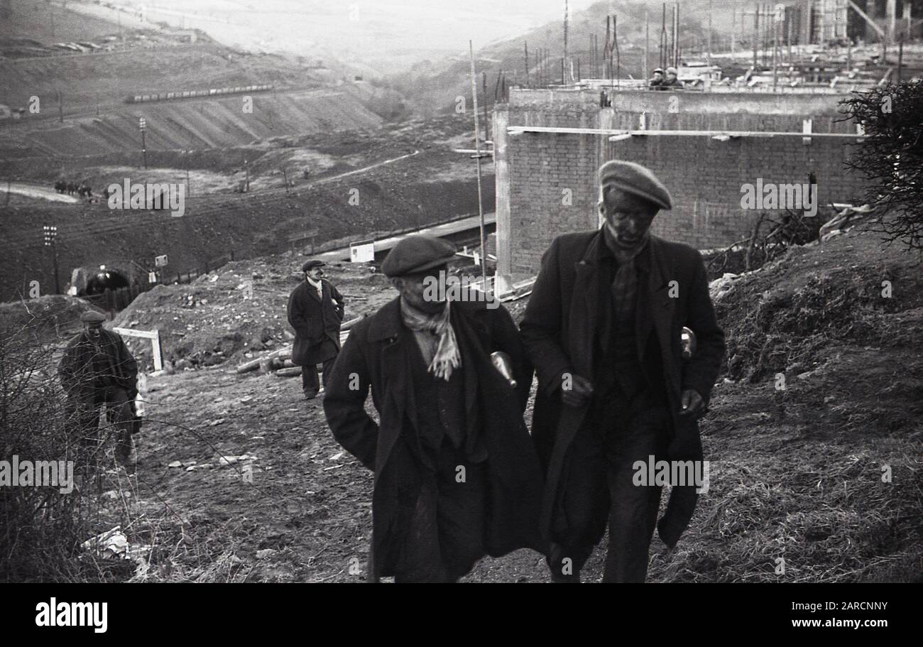 1940s, storico, lavoratori delle miniere indossando soprabiti e cappotti e con facce annerite coperte di fuliggine, camminando sulla collina dalla miniera di carbone nella valle sottostante, Merthyr, Galles, Regno Unito. Foto Stock