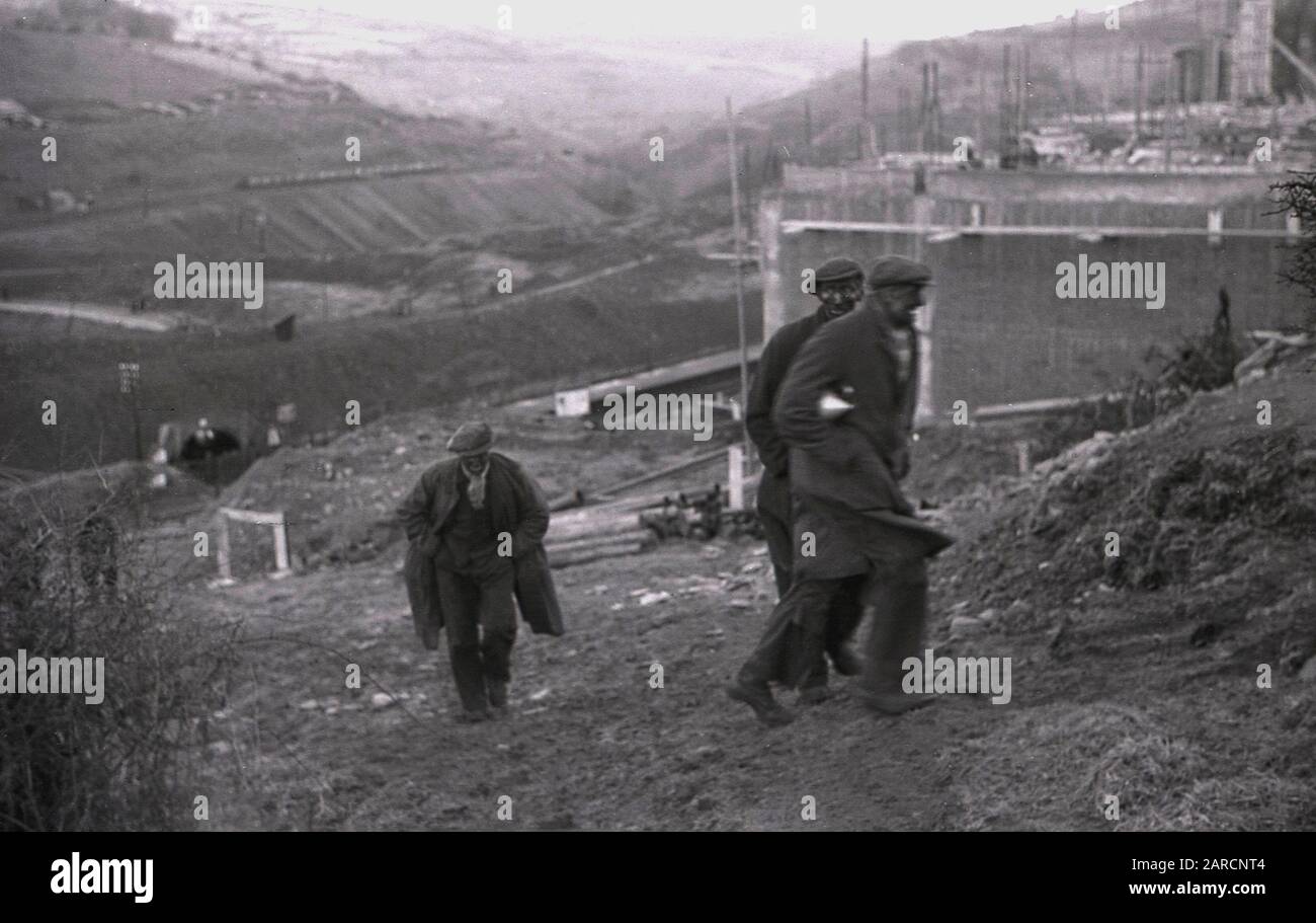 1940s, storico, lavoratori delle miniere indossando soprabiti e cappotti e con facce annerite coperte di fuliggine, camminando sulla collina dalla miniera di carbone nella valle sottostante, Merthyr, Galles, Regno Unito. Foto Stock