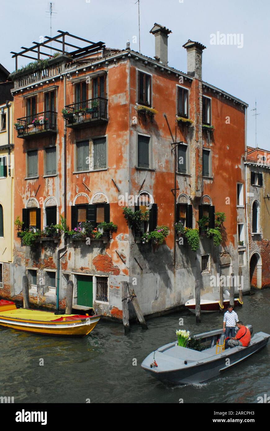 Una scena su un canale a Venezia, in Italia, con barche che vanno in giro per il business quotidiano. Un uomo trasporta un mazzo di gigli su una barca. Foto Stock