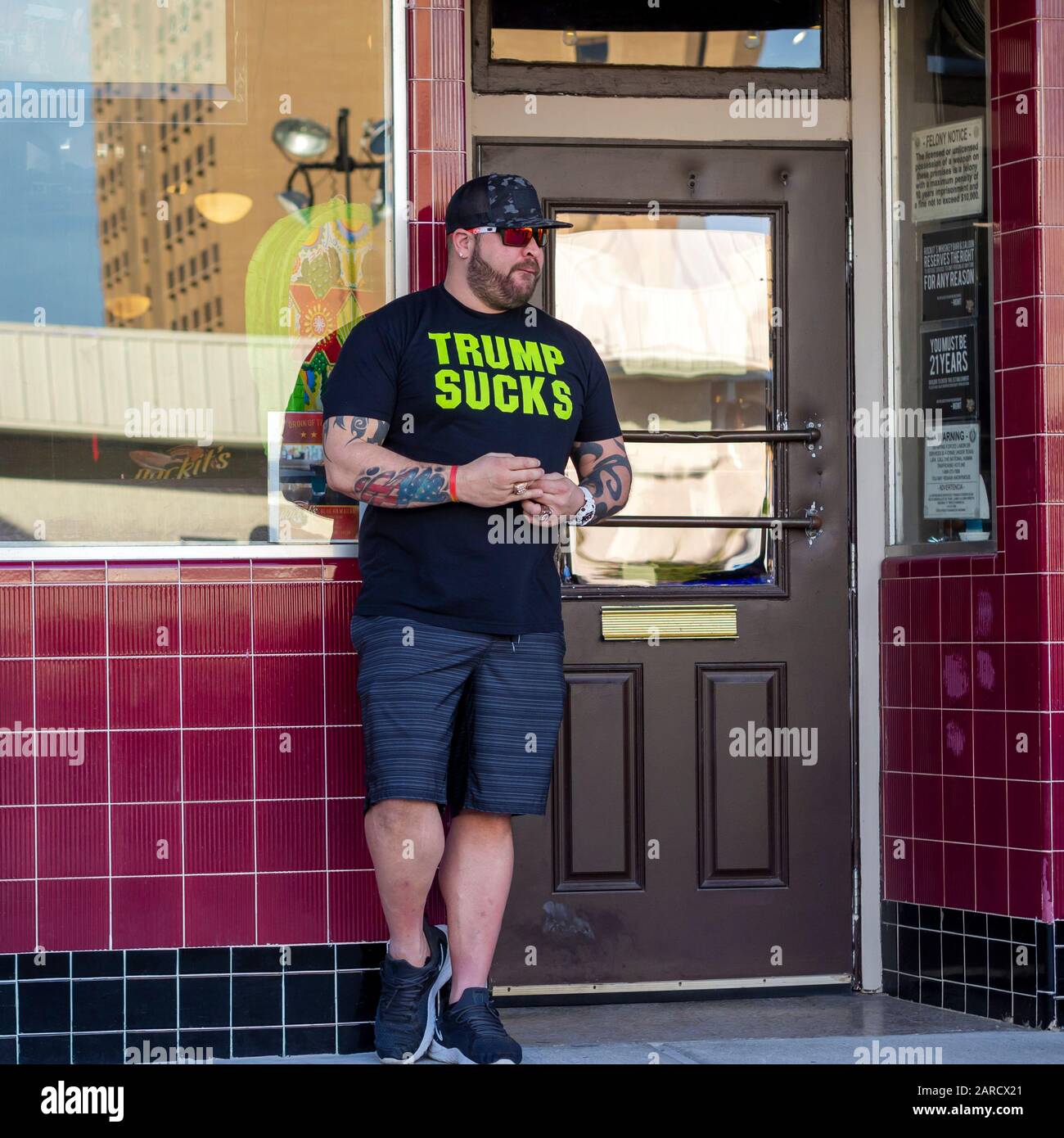 Il potente bouncer con le braccia tatuate indossa una t-shirt nera con "Trump Succhia" in grandi lettere verdi alla porta di un bar a Corpus Christi, Texas USA. Foto Stock