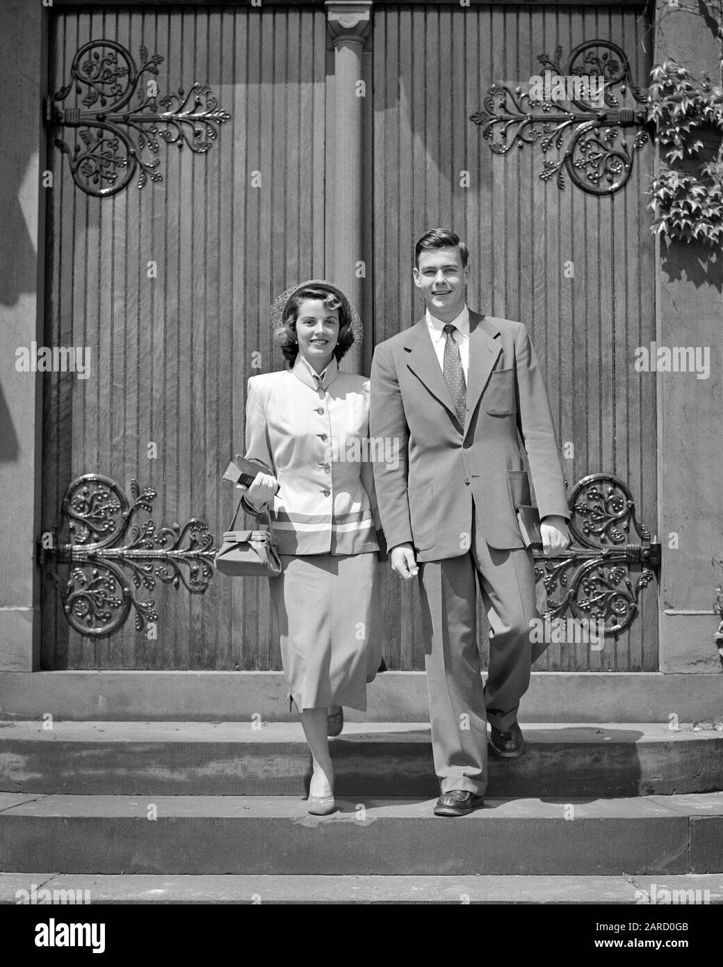 1950S UOMO DONNA GIOVANE COPPIA CHE CAMMINA FUORI DALLA CHIESA VESTITO DI  DOMENICA MIGLIORI VESTITI SORRIDENTE GUARDANDO LA MACCHINA FOTOGRAFICA -  C3399 HAR001 HARS GIOVANE ADULTO EQUILIBRIO LAVORO DI SQUADRA MEGLIO FELICE