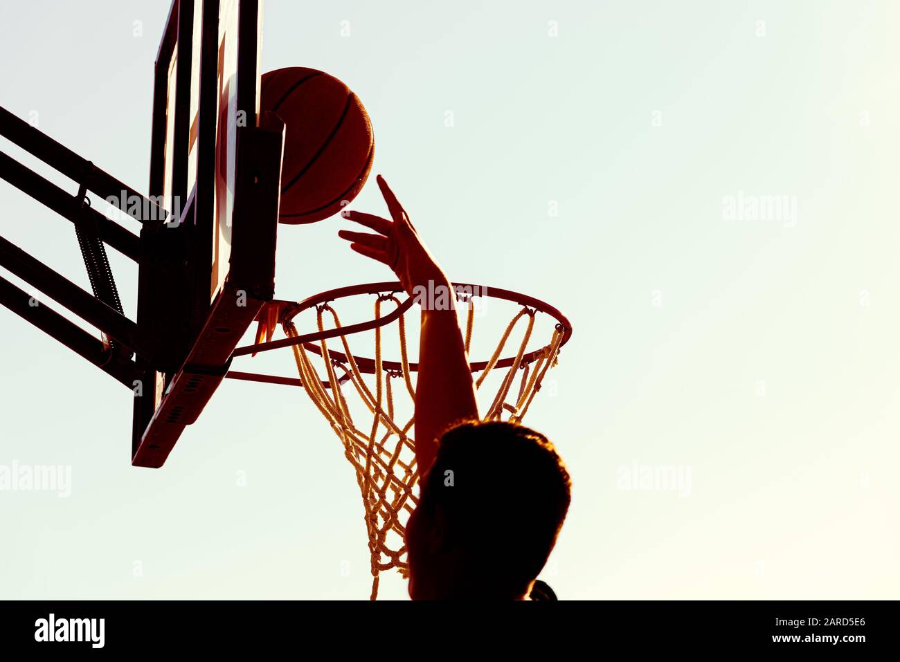 Il giovane giocatore di basket dalla silhouette sinua che salta in alto sul cerchio di basket raggiunge un punteggio con un dunk di volo al tramonto. Successo e obiettivo Foto Stock