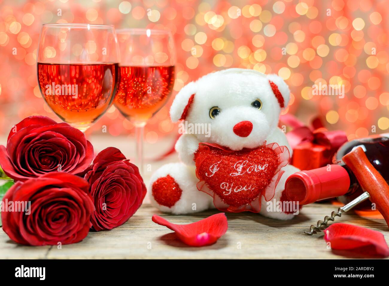 Orso peluche bianco giocattolo, rose rosse, scatola regalo, bottiglia di vino, cavatappi e bicchieri di vino su un vecchio tavolo di legno di fronte a uno sfondo luminoso sfocato. Foto Stock