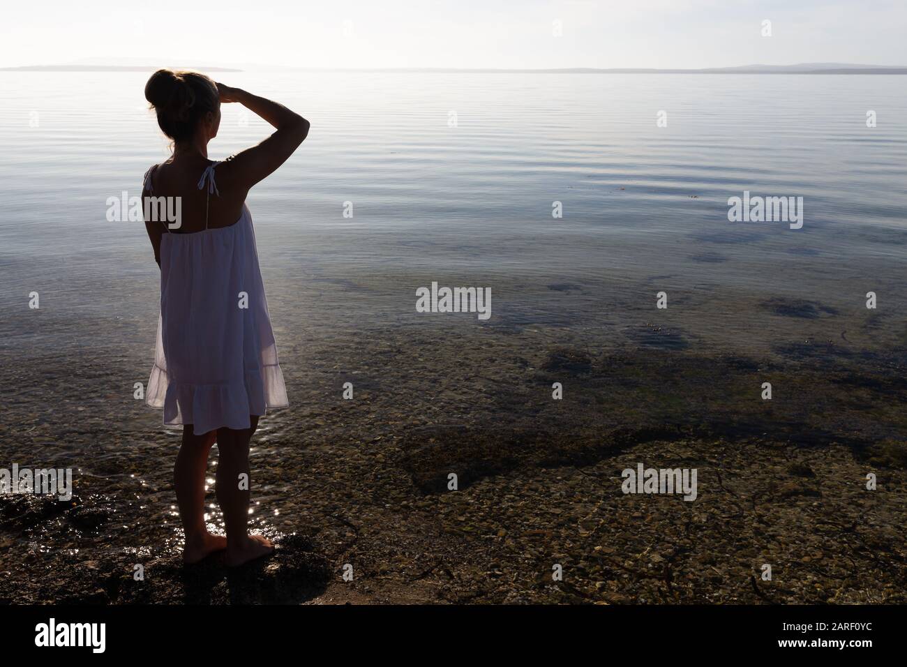 Bella giovane donna in abito bianco con vista sulla splendida e incontaminata baia costiera su una tranquilla mattinata in Australia meridionale. Foto Stock