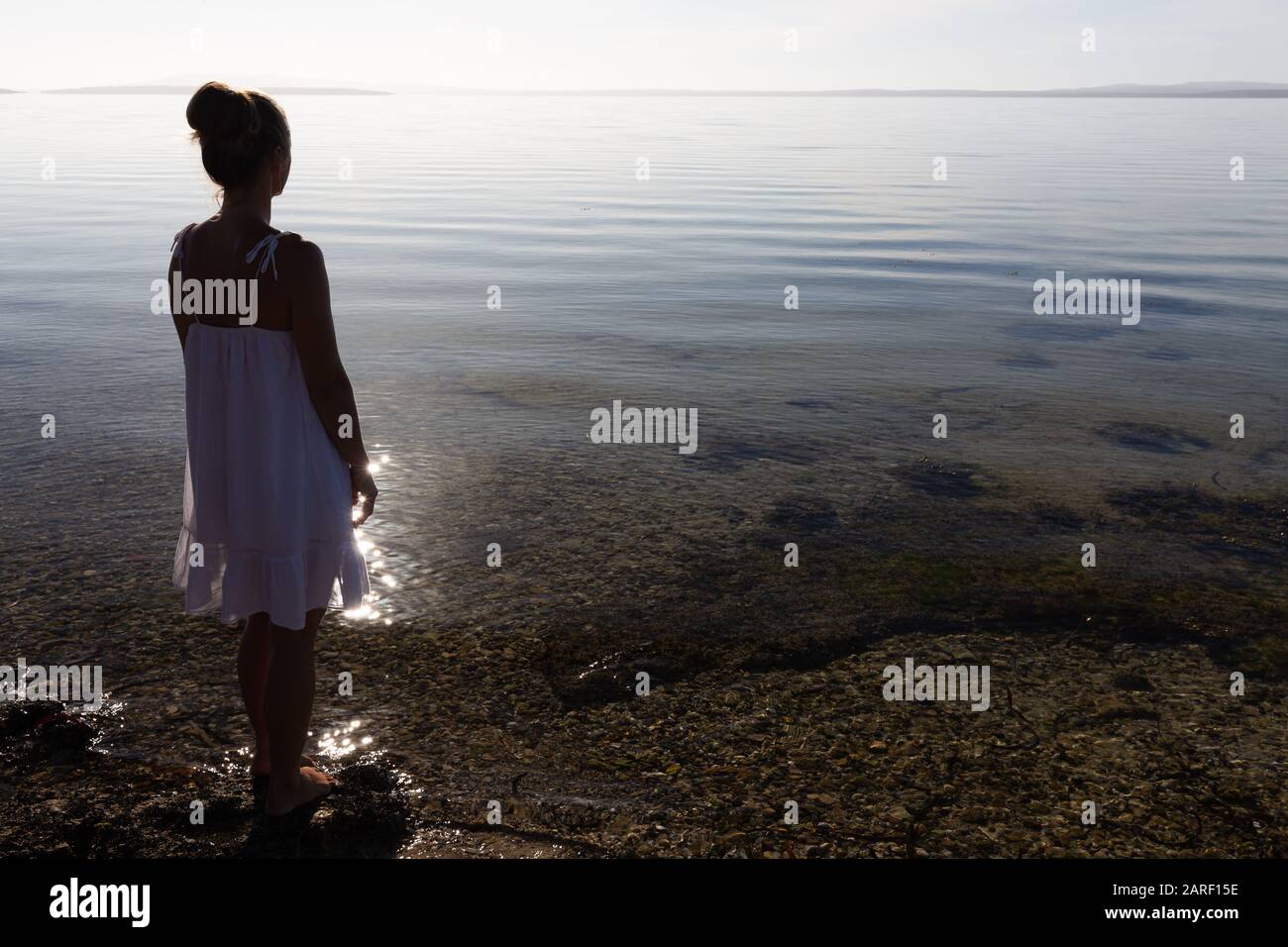 Donna giovane di colore bianco, con silhouette e luce sul retro, con vista sulla splendida e incontaminata baia costiera, in una tranquilla mattinata in South Australia. Foto Stock