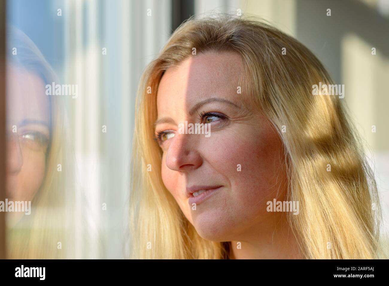 Attraente donna bionda in piedi sognando guardando longingly fuori attraverso una finestra riflessa nel vetro Foto Stock