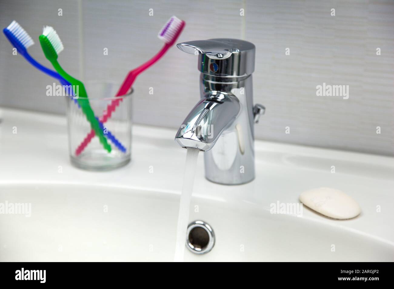 Spazzolini da denti, sapone e rubinetto d'acqua con acqua corrente pulita in un lavandino bianco da bagno. Foto Stock