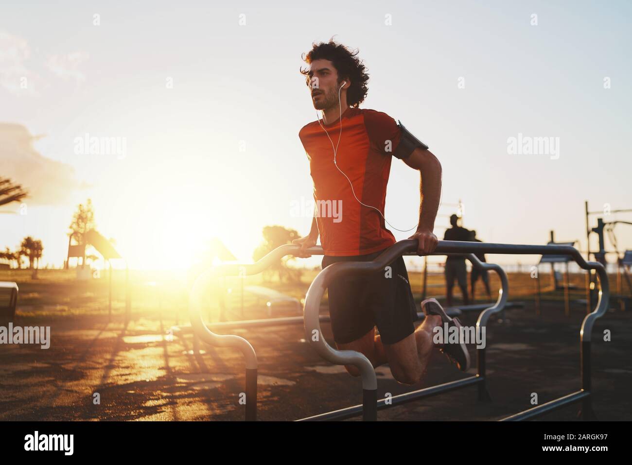 Atletica giovane atleta maschile ascoltare musica su auricolari esercizio su bar paralleli fare push-up nel parco - giovane uomo facendo dips all'aperto Foto Stock
