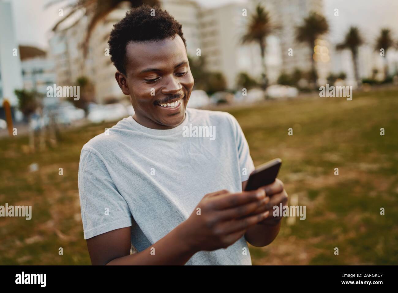 Ritratto di un giovane atleta di sesso maschile che si alza al parco sorridendo mentre messaggi di testo sul telefono cellulare Foto Stock