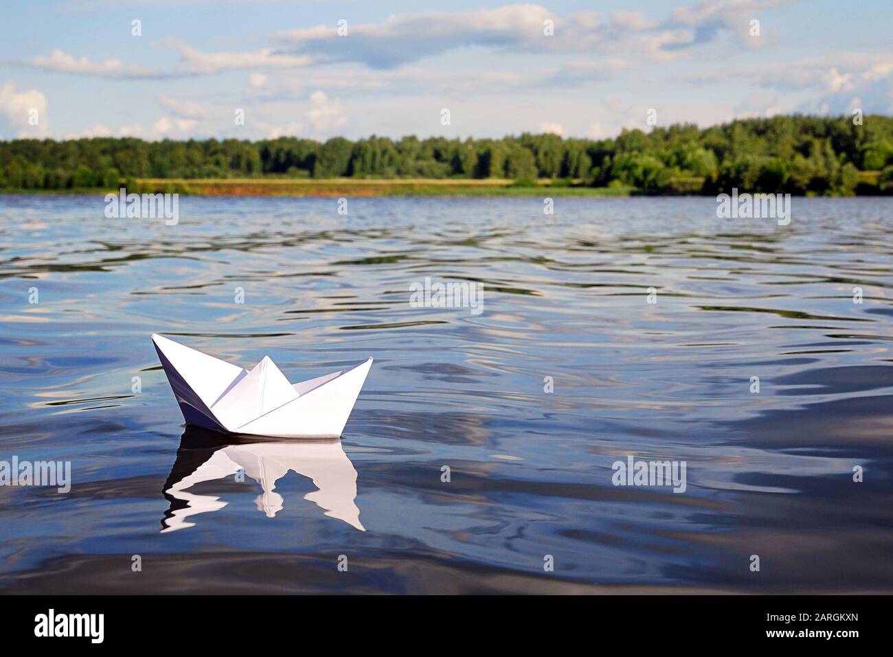 Piccola barca di carta che galleggia su acque blu calmo del fiume, foresta verde, cielo blu con le nuvole estive chiare sull'orizzonte. Libertà, avventura, sogno e tra Foto Stock