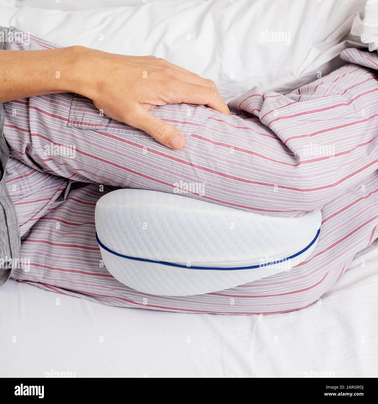 vestirsi di un uomo caucasico, indossando pantaloni pigiama a righe, usando un cuscino anatomico tra le gambe mentre è sdraiato su un letto con lenzuola bianche Foto Stock