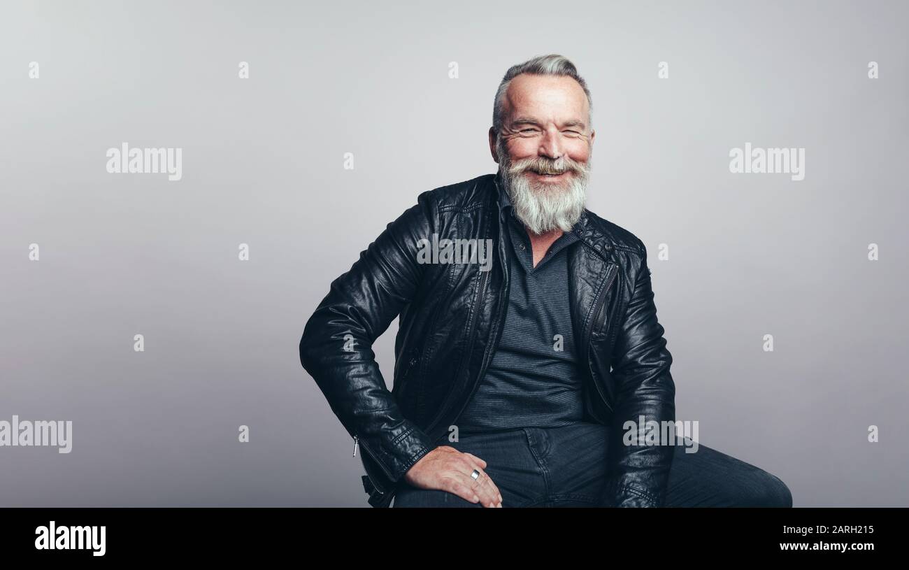 Uomo anziano con barba bianca che guarda la fotocamera. Uomo sorridente in giacca nera seduta su sfondo grigio. Foto Stock