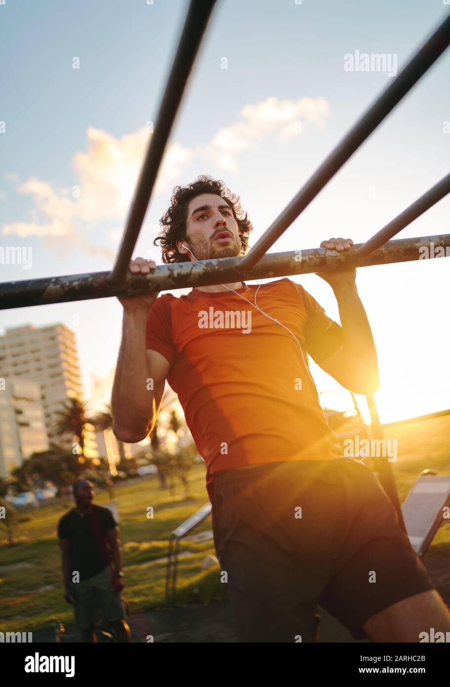 Indossate un giovane con gli auricolari nelle orecchie allenando i muscoli delle braccia sui bar della palestra all'aperto in estate - uomo che fa pullup all'aperto Foto Stock