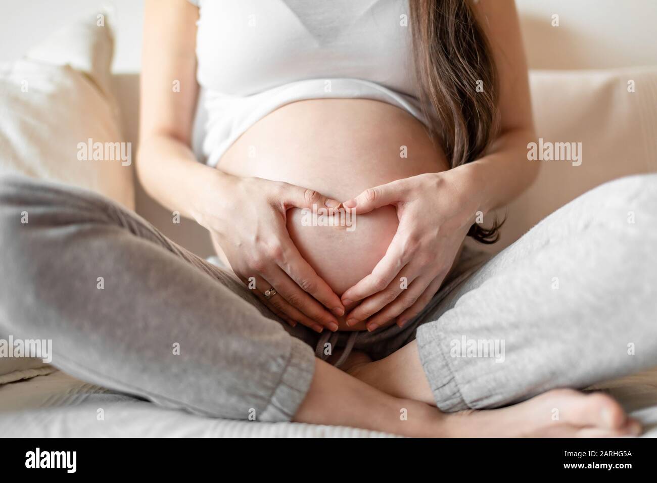 Giovane donna incinta seduta su divano bianco e tenendo le mani sul ventre Foto Stock