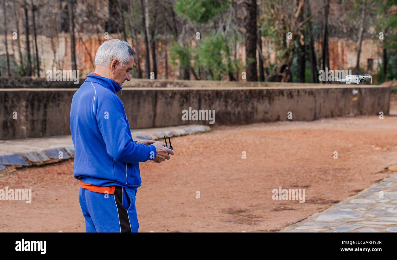 Vista laterale del vecchio uomo che vola il drone in abbigliamento sportivo mentre si trova contro gli alberi nel parco Foto Stock
