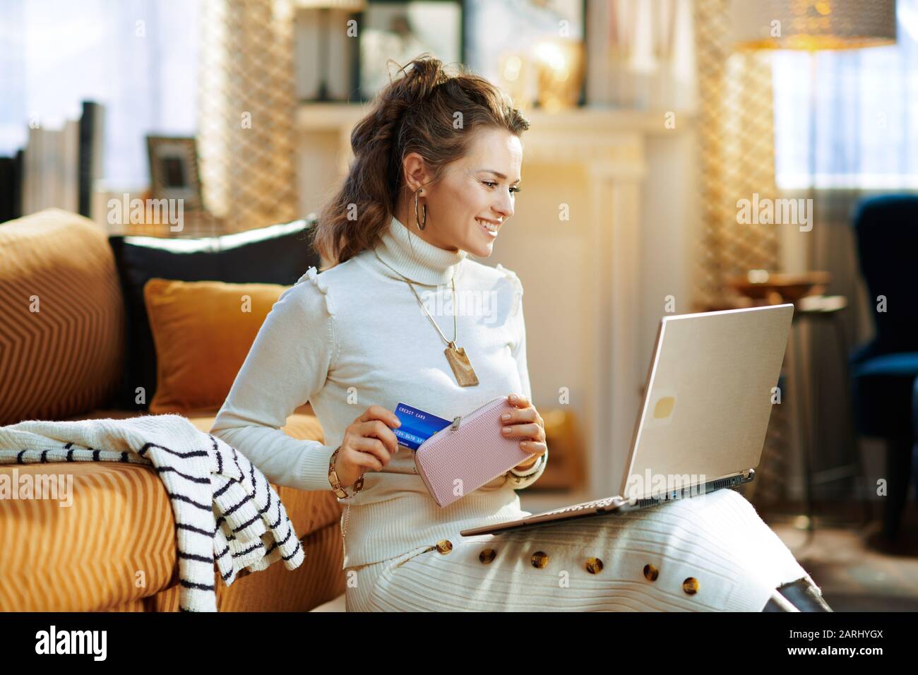 felice donna di mezza età in maglione bianco e gonna con carta di credito seduta vicino divano alla ricerca di nuovo maglione al negozio online moda su un computer portatile in th Foto Stock