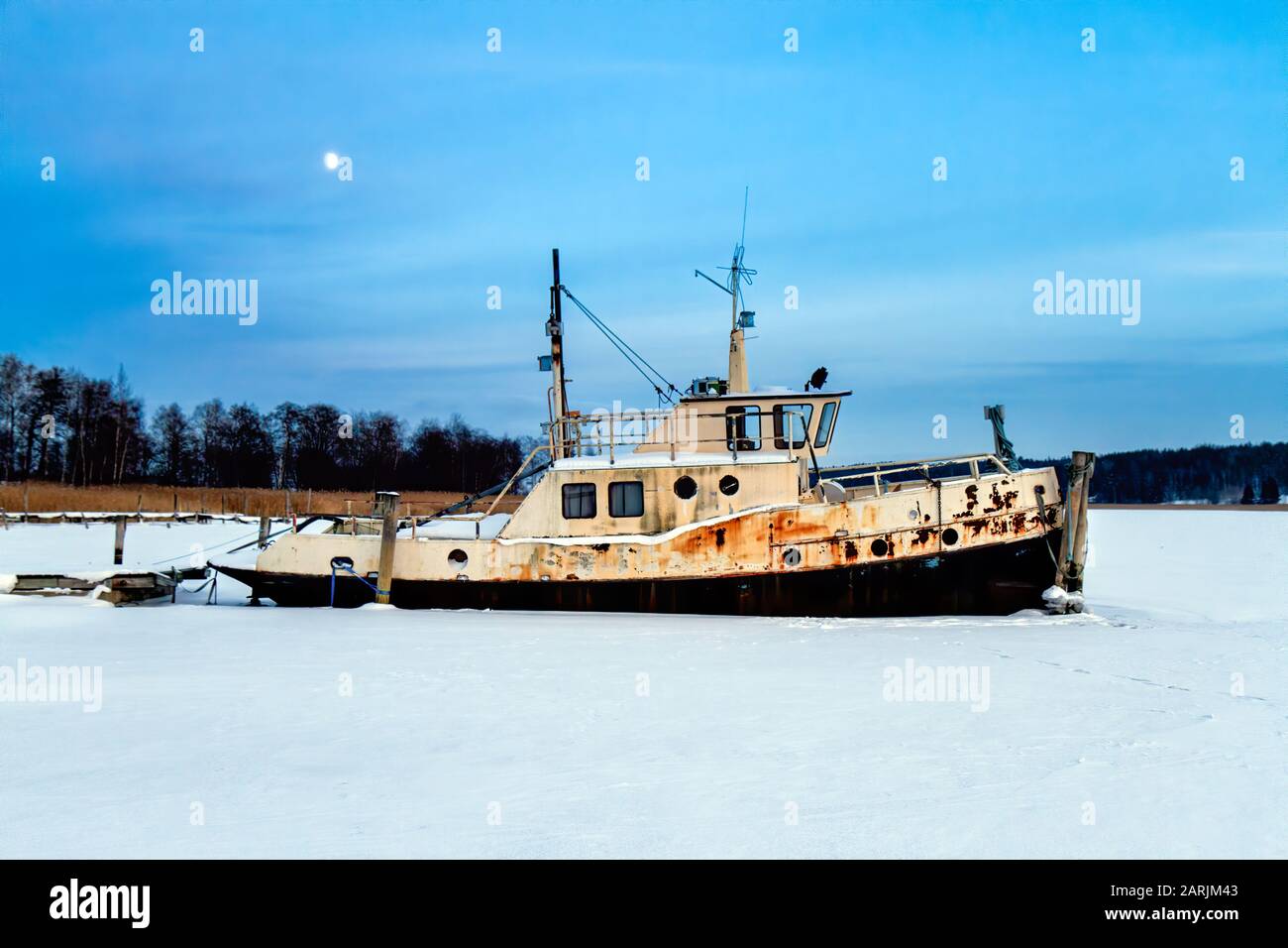 Antico rimorchiatore piccolo e arrugginito al molo in una fredda serata invernale. Barca bloccata in ghiaccio, Kaarina, Finlandia. Foto Stock