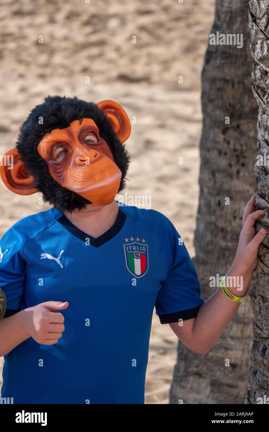 Giovane ragazzo con camicia da calcio Blur Italia che indossa una maschera di scimmia e che si appoggia verso una palma su una spiaggia. Foto Stock