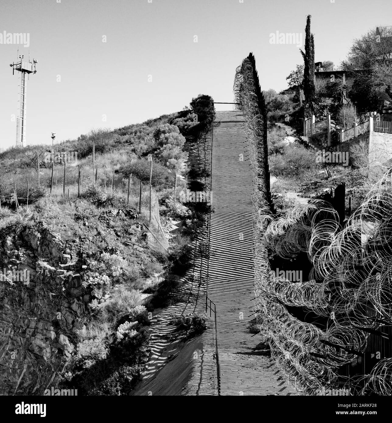 Vista della recinzione di confine internazionale USA/Messico con filo di rasoio in salita con le case messicane sull'altro lato della recinzione da Nogales, AZ, USA, B&W. Foto Stock