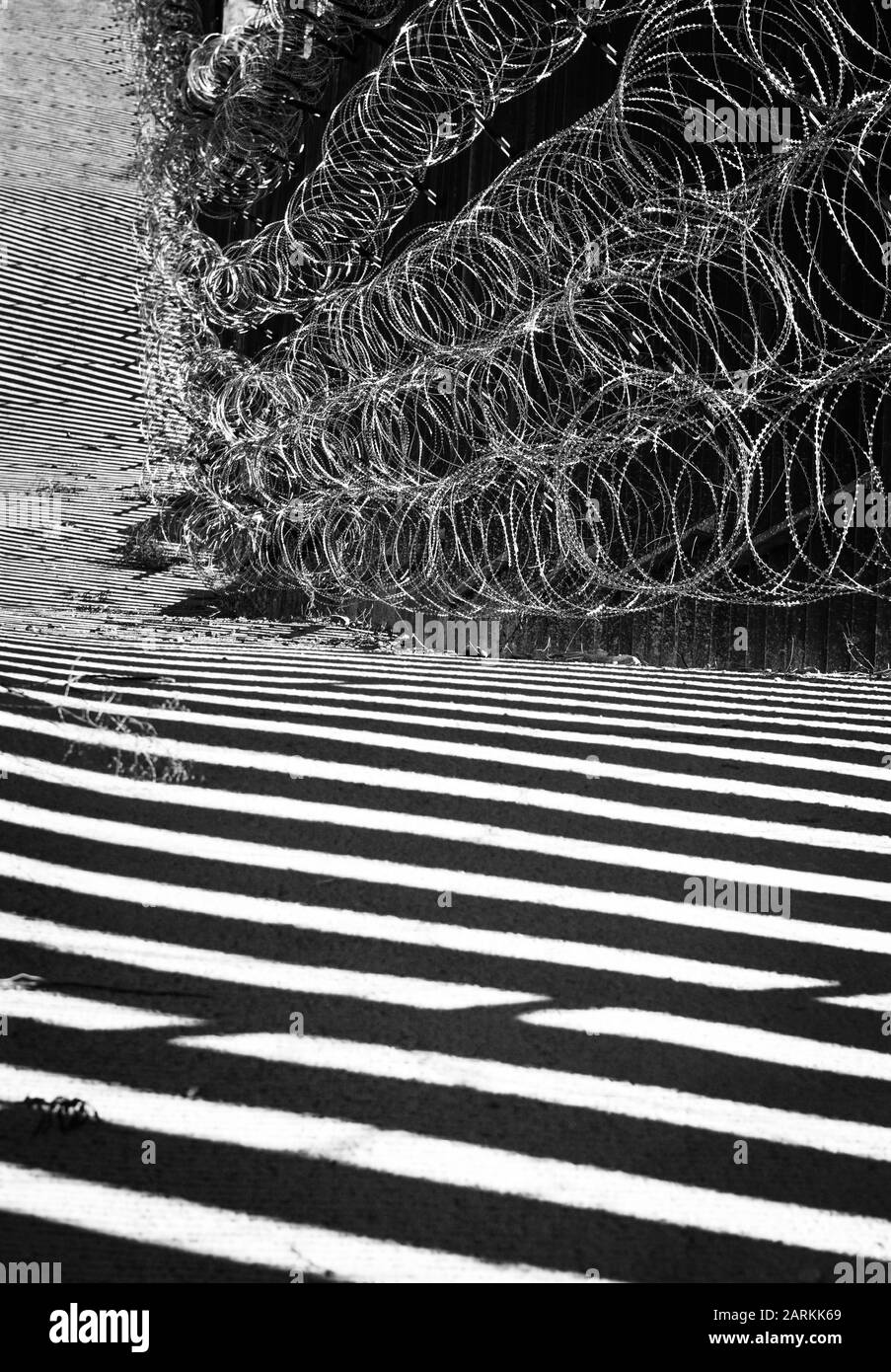 Vista astratta della recinzione di confine internazionale USA/Messico con filo di rasoio con luce e ombre drammatiche a Nogales, AZ, USA, in bianco e nero Foto Stock