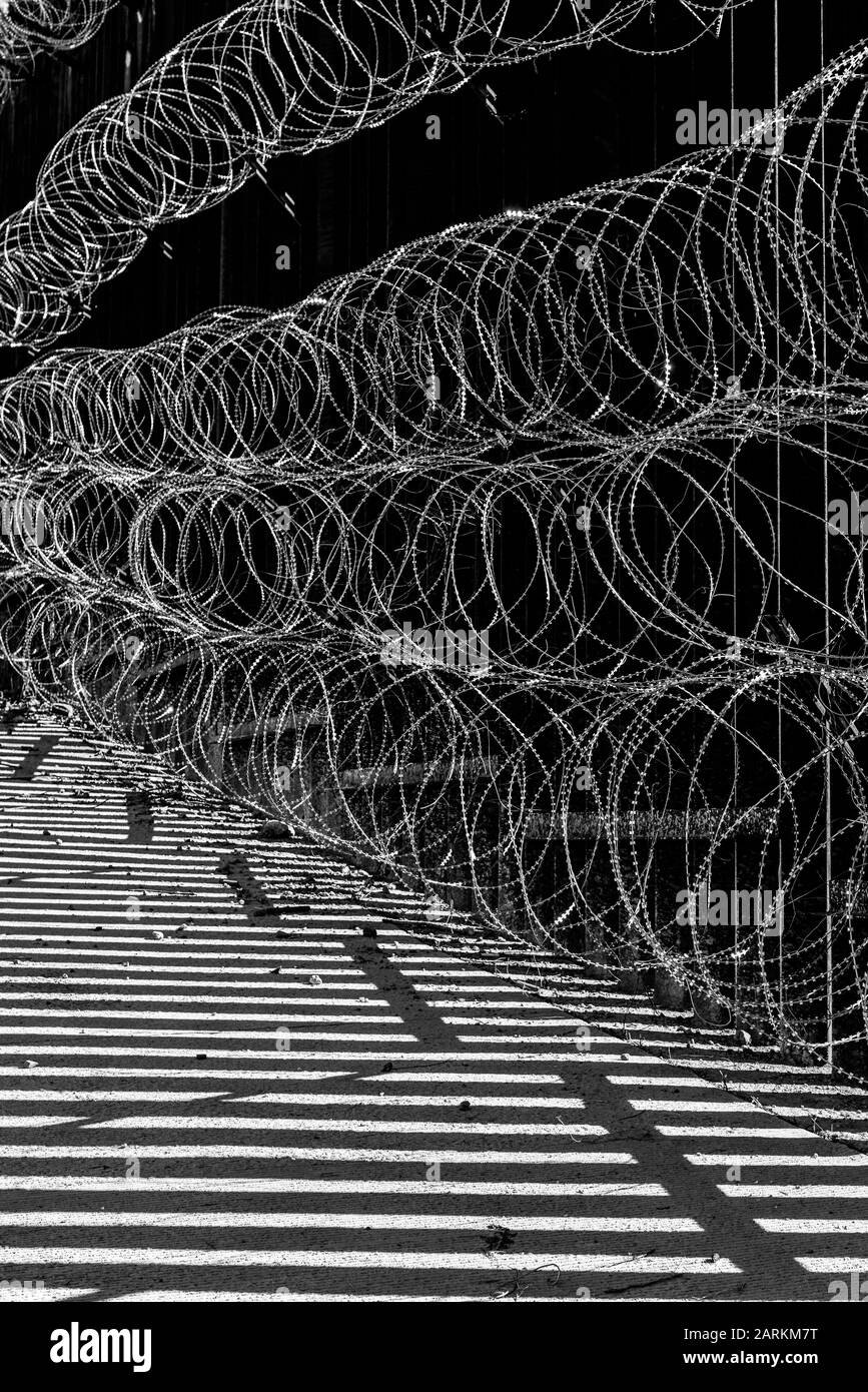 Vista astratta della recinzione di confine internazionale USA/Messico con filo di rasoio con luce e ombre drammatiche a Nogales, AZ, USA, in bianco e nero Foto Stock