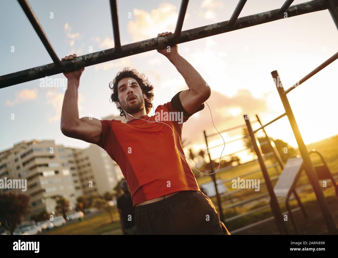 Ritratto di un giovane uomo muscoloso che ascolta musica sulle cuffie che lavorano fuori sulle barre della scimmia facendo pull-up in palestra all'aperto Foto Stock