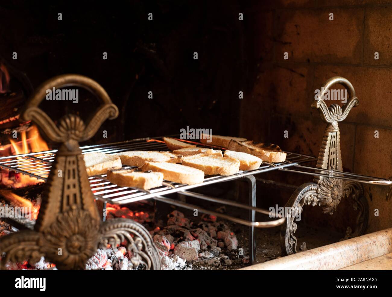 Pane affettato assortito tostare su una rastrelliera metallica su carboni caldi e fuoco con fiamme in un camino all'interno con spazio copia sopra Foto Stock
