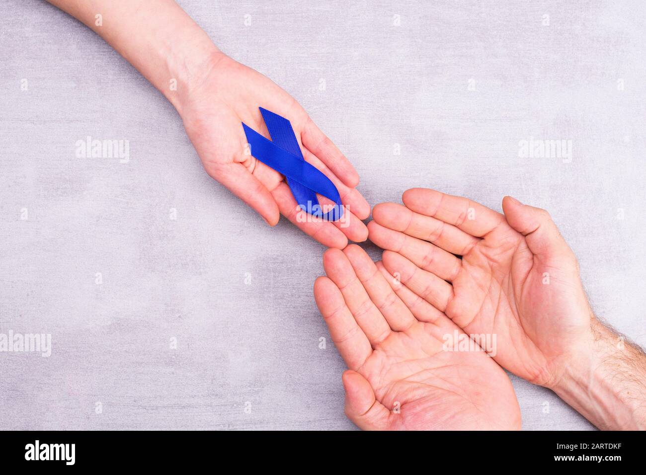 Una donna tiene fuori un nastro blu di consapevolezza, simbolo nelle mani maschili, cancro del colon, cancro colorettale su uno sfondo grigio con spazio per il testo, piatto lay Foto Stock