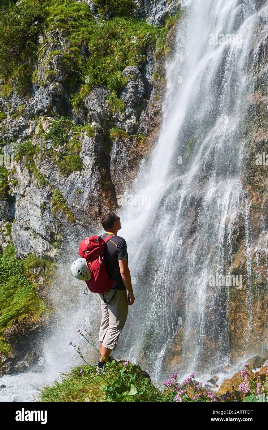 Uomo con casco bianco sul suo bacpack rosso che guarda verso la cascata Dalfazer (Dalfazer Wasserfall) sopra Achensee, Austria, in una luminosa giornata di sole. Foto Stock