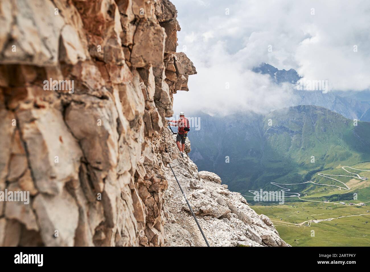 Uomo in via ferrata Cesare Piazzetta, Dolomiti, Italia, vicino alle pareti rocciose, con una tortuosa strada sottostante, in una giornata estiva con nuvole Foto Stock