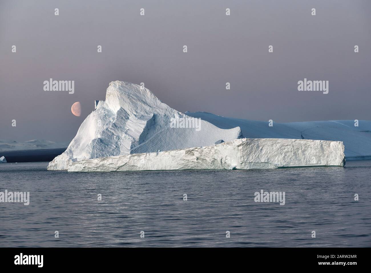 Enorme iceberg galleggiante in un fiordo al tramonto con la luna sulla sinistra. Scoresby Sund, Kangertitivaq, Groenlandia, Danimarca Foto Stock