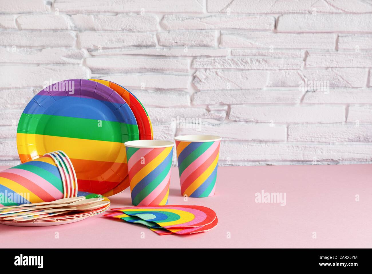 Vasellame ecologico a colori arcobaleno su sfondo rosa su pareti in mattoni bianchi. Accessori per feste di compleanno, concetto di utensile da picnic. Spazio di copia Foto Stock