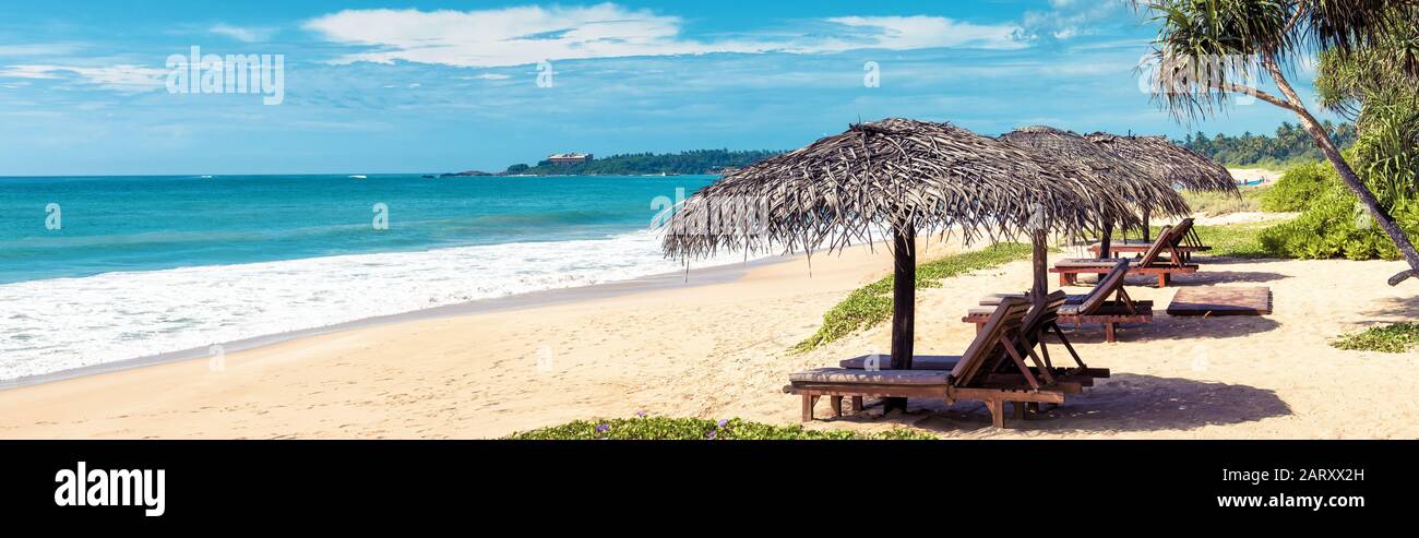 Lettini da spiaggia con ombrelloni sulla spiaggia tropicale. Coniglietto orizzontale per intestazione sito web. Vista panoramica su una spiaggia di sabbia soleggiata in tropico. Beautifu Foto Stock