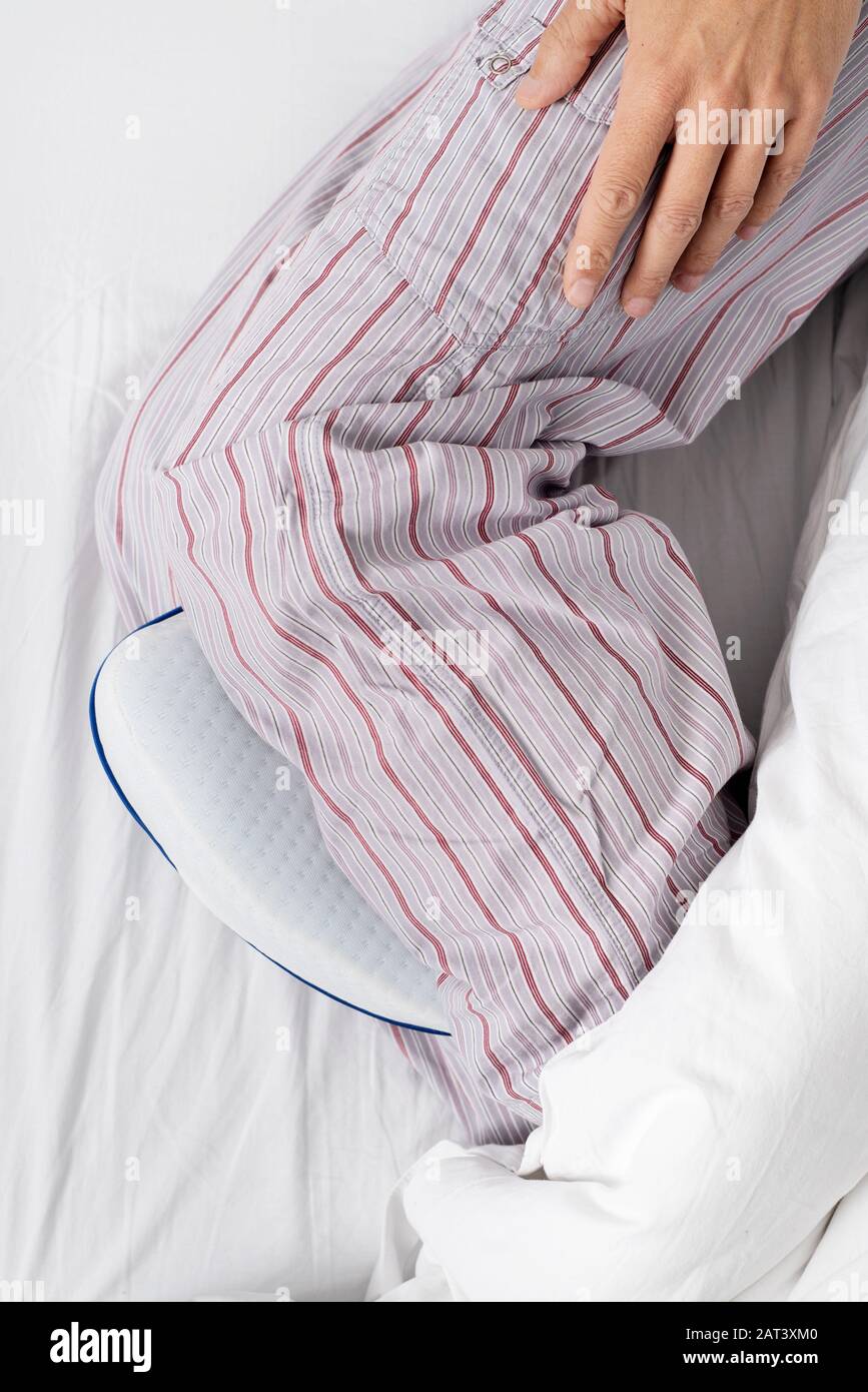 vista ad alto angolo di un uomo caucasico, indossando pantaloni pigiama a righe, utilizzando un cuscino anatomico tra le gambe mentre è sdraiato su un letto con set di bianco Foto Stock