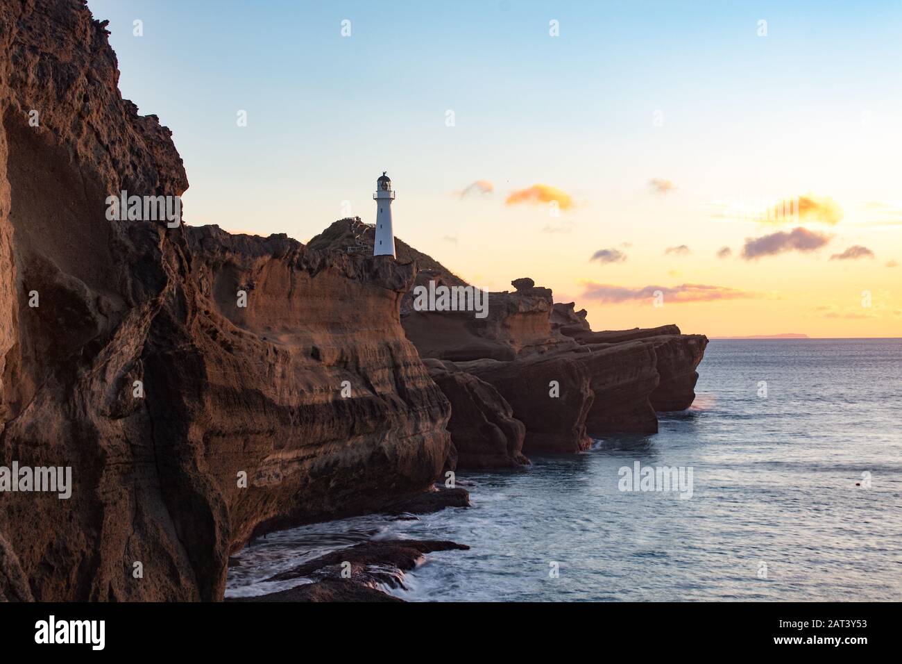 Vista sul faro di Castlepoint in piedi su enormi scogliere scoscese sopra l'oceano durante il tramonto arancione con poche nuvole. Nuova Zelanda, Isola Del Nord Foto Stock