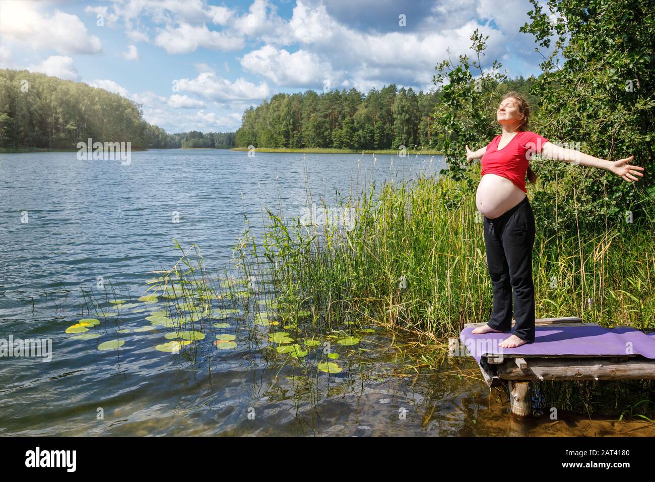 Giovane donna incinta levigare su un molo di legno in un lago di foresta durante la calda giornata di sole spargendo le mani godendo della natura - sano lifes gravidanza Foto Stock