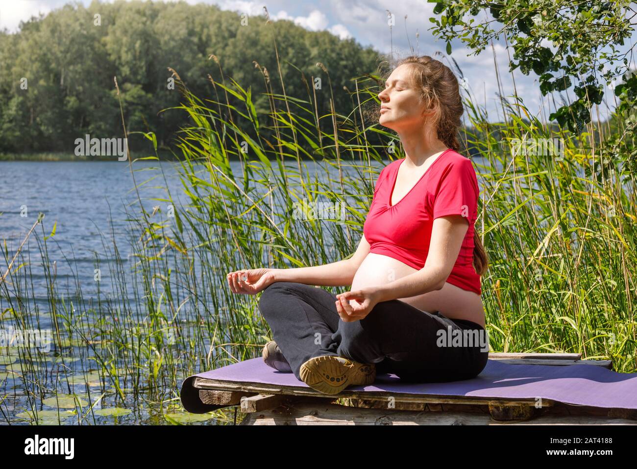 Giovane donna incinta vestita sportivo fare esercizio di yoga seduto in posizione lotus su un molo di legno in un lago di foresta meditando. Fitness e guarire Foto Stock
