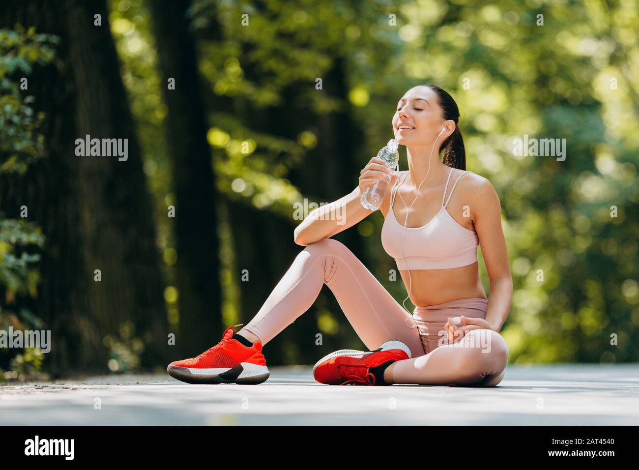 Una donna bruna beve un'acqua negli auricolari seduti nel parco all'aperto - immagine Foto Stock