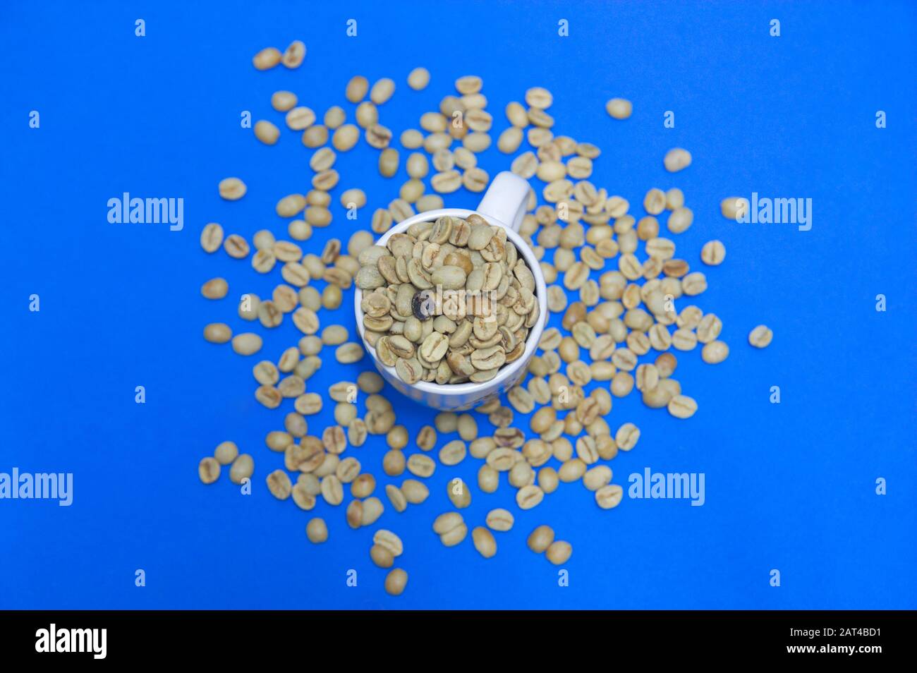 Una tazza di caffè pieno di chicchi di caffè crudi e non tostati visti dall'alto. Inoltre, i chicchi di caffè sono anche sparsi sul tavolo che è blu classico Foto Stock